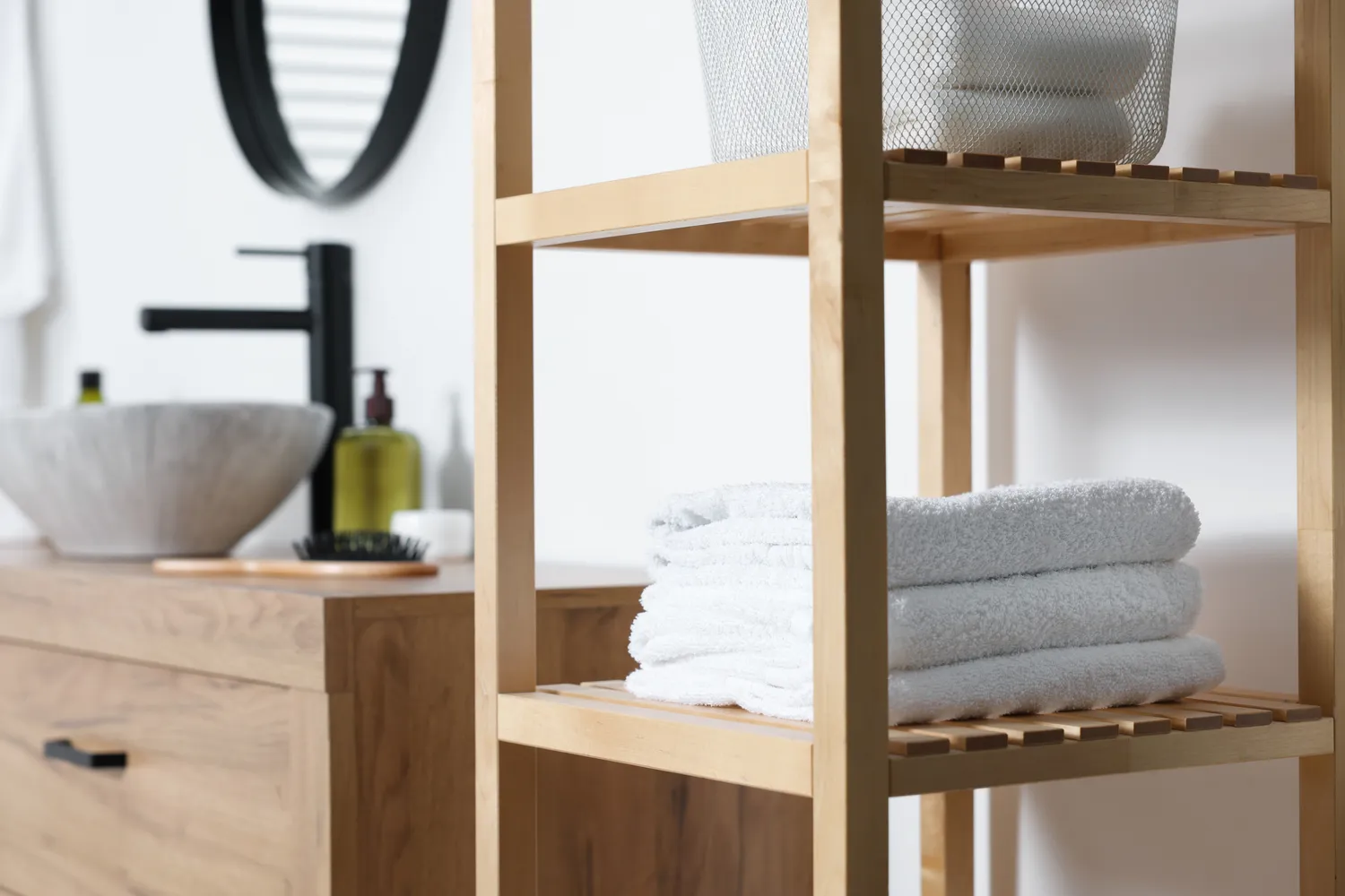 Antjes – Roomstyling – Ein modernes Badezimmer mit einem Holzregal, auf dem gefaltete weiße Handtücher liegen, neben einem Waschtisch mit Steinwaschbecken und schwarzem Wasserhahn.