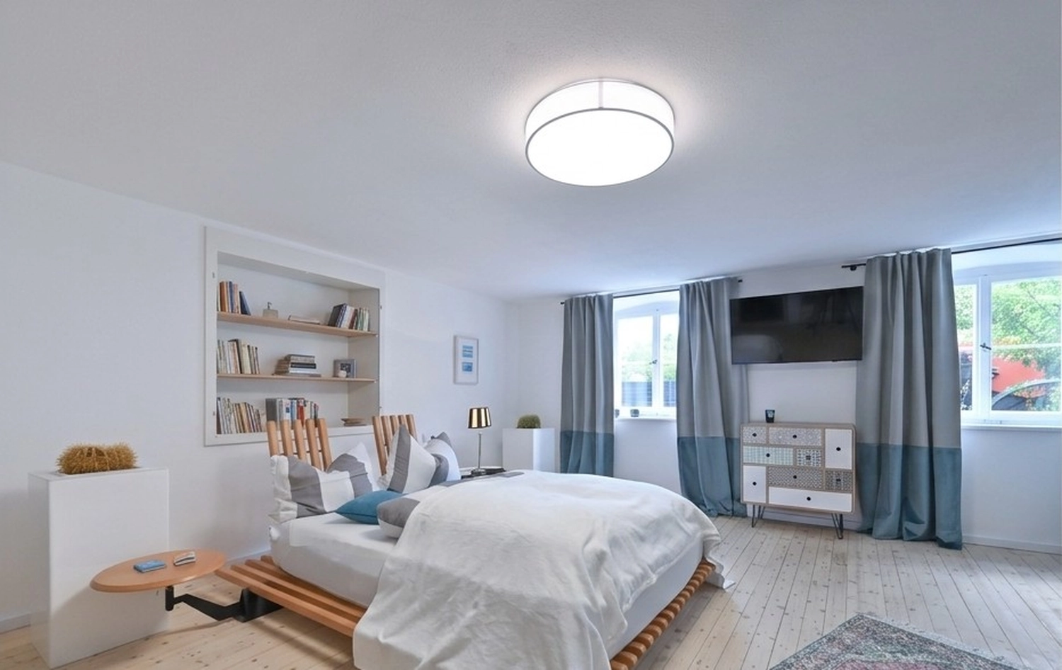 Antjes – Roomstyling – Ein helles, minimalistisches Schlafzimmer mit Holzbett, weißer Bettwäsche, eingebautem Bücherregal und blaugrauen Vorhängen.
